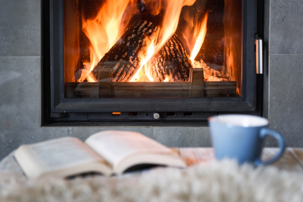 Open book, cup of tea and warm plaid near burning fireplace. Hygge concept